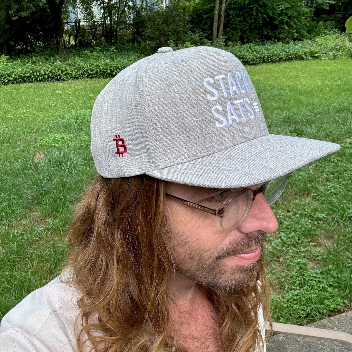 A model with long hair wears a heather grey hat with the words 'STACK SATS' embroidered on the front in white. On the side is a bitcoin logo embroidered in red. He's standing outside on a green lawn.