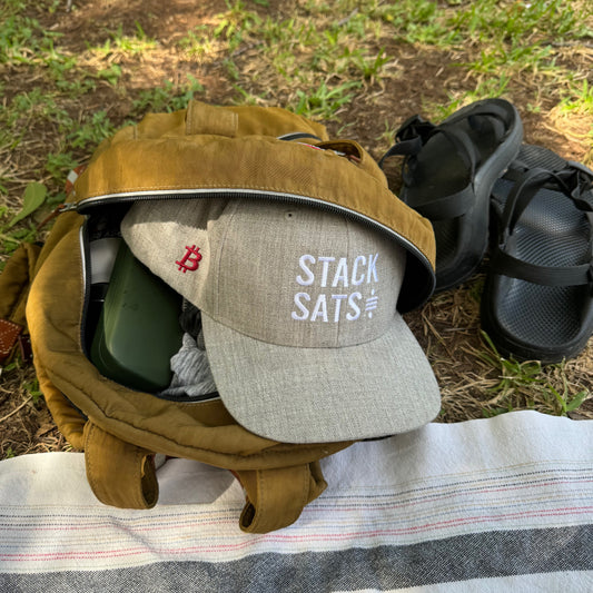 A heather grey hat with the words 'STACK SATS' embroidered on the front in white and a bitcoin logo embroidered in red on the side, sits partly emerging from a tan backpack sitting on a green lawn next to sandals and a beach towel.