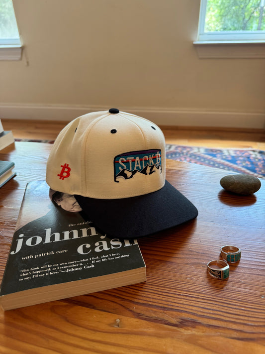 A white and black baseball cap with a blue and red mountain logo that says 'STACK Bitcoin' on the front. The cap has an orange Bitcoin symbol on the side. The cap is sitting on top of a book titled 'johnny cash the autobiography'. There are two silver rings with turquoise stones on a wooden surface.