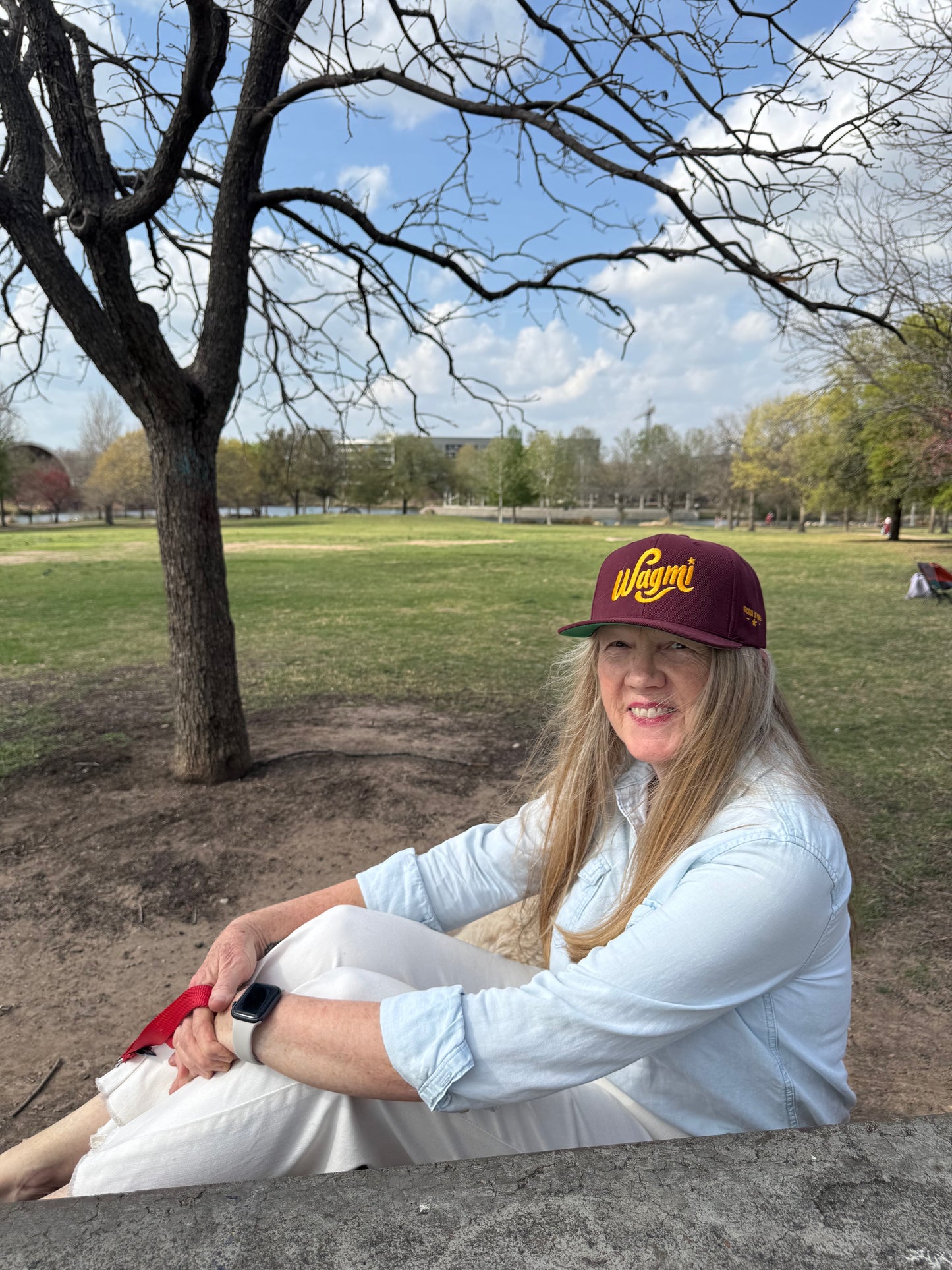 A female model sits on a park bench wearing a maroon baseball cap that reads WAGMI (We’re All Gonna Make It) embroidered in yellow, and 'Bitcoin is hope' on the side.