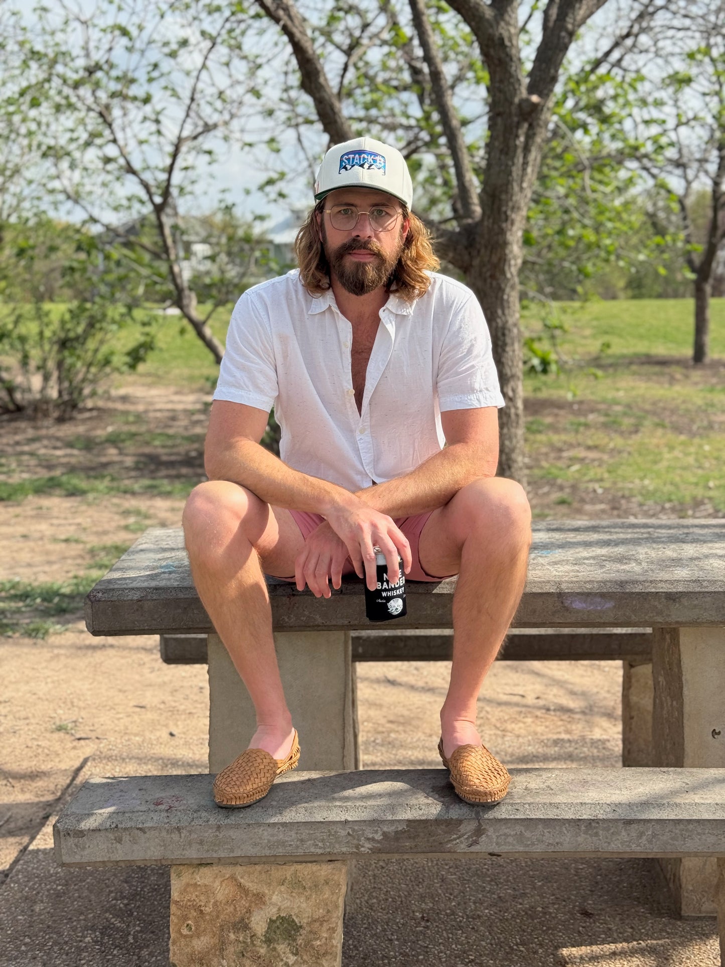 Model sits on a park bench wearing leather shoes and holding a beer. He wears a natural and black billed trucker hat that features embroidered mountains and the text "Stack" with the bitcoin logo.