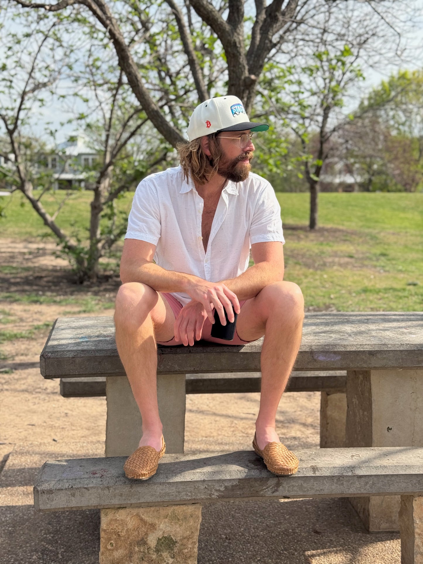 Model sits on a park bench wearing leather shoes and holding a beer. He wears a natural and black billed trucker hat that features embroidered mountains and the text "Stack" with the bitcoin logo. He's looking to his left to reveal an orange Bitcoin logo embroidered on the side of the hat.