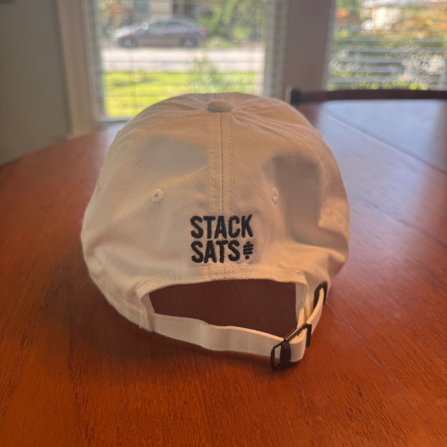 A white dad hat with 'STACK SATS' embroidered in black on the back lays on a mid century modern dining table.