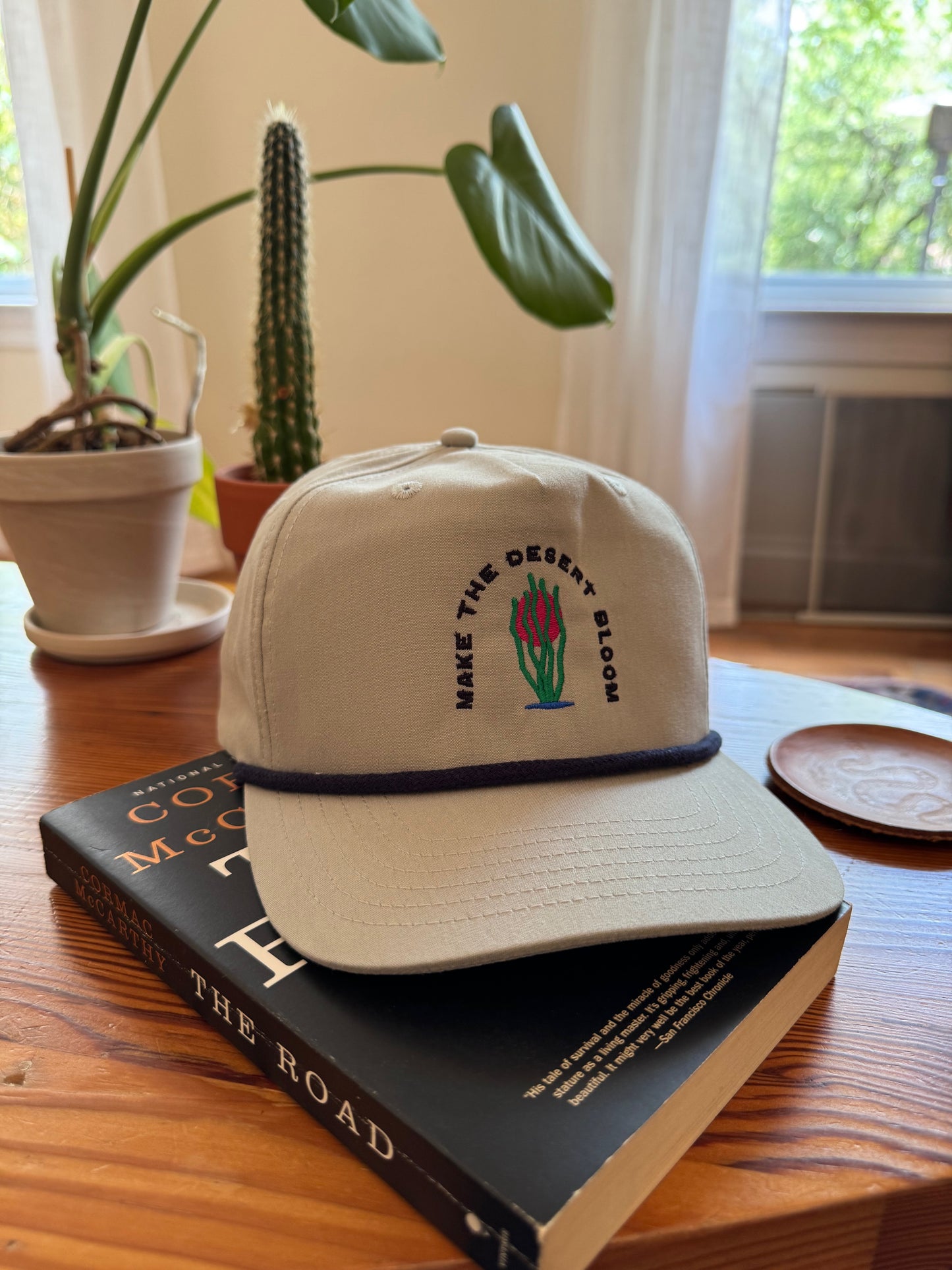 A grey golf rope cap with a navy rope sits on a wooden coffee table on top of a book called "The Road." Embroidered on the hat is an ocotillo in front of a setting sun and under the words "Make the desert bloom."