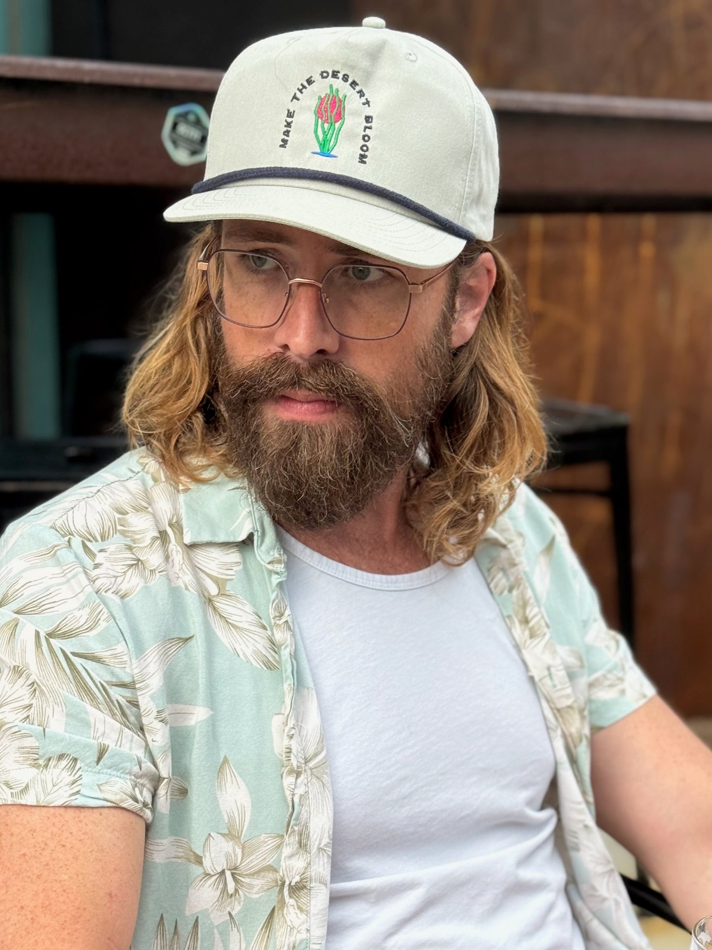 Man with a beard wearing a grey golf rope cap with a navy rope. Embroidered on the hat is an ocotillo in front of a setting sun and under the words "Make the desert bloom."