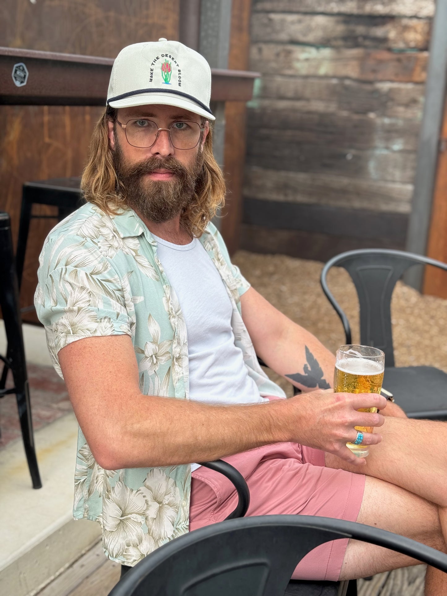 A model holding a beer wears a grey golf rope cap with a navy rope. He looks directly into camera. Embroidered on the hat is an ocotillo in front of a setting sun and under the words "Make the desert bloom."