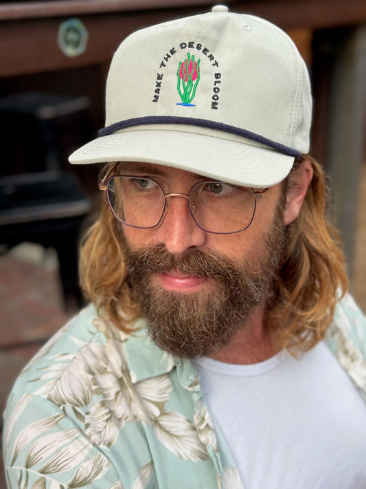 A model with a beard and long hair wears a grey golf rope cap with a navy rope. Embroidered on the hat is an ocotillo in front of a setting sun and under the words "Make the desert bloom."