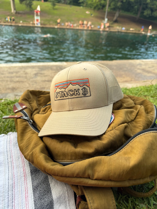 Khaki Bitcoin Stack Mountain Retro Trucker Hat with embroidered mountain design, sitting by a pool on a backpack, perfect for crypto enthusiasts.