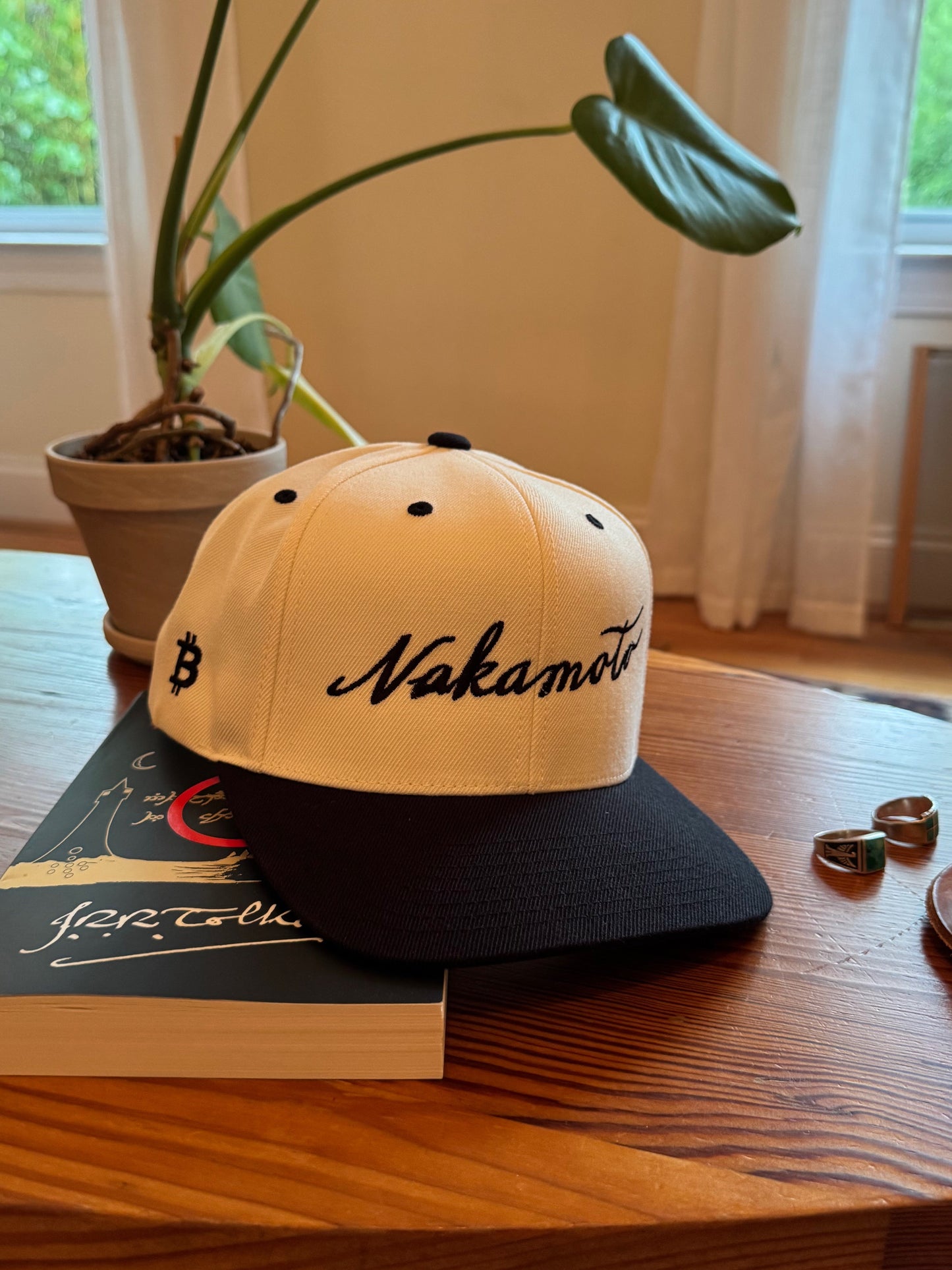 A beige and black trucker hat with the word 'Nakamoto' embroidered in a script font on the front in black, and a bitcoin logo embroidered in black on the side. The hat sits on a book that says 'J.R.R. Tolkien' in a script font, and next to two turquoise rings on a wooden coffee table.