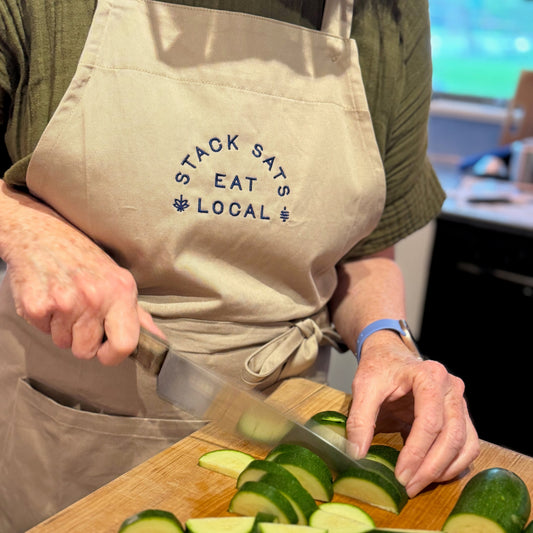 Person in a 'Stack Sats Eat Local' apron slicing cucumbers on a wooden cutting board.