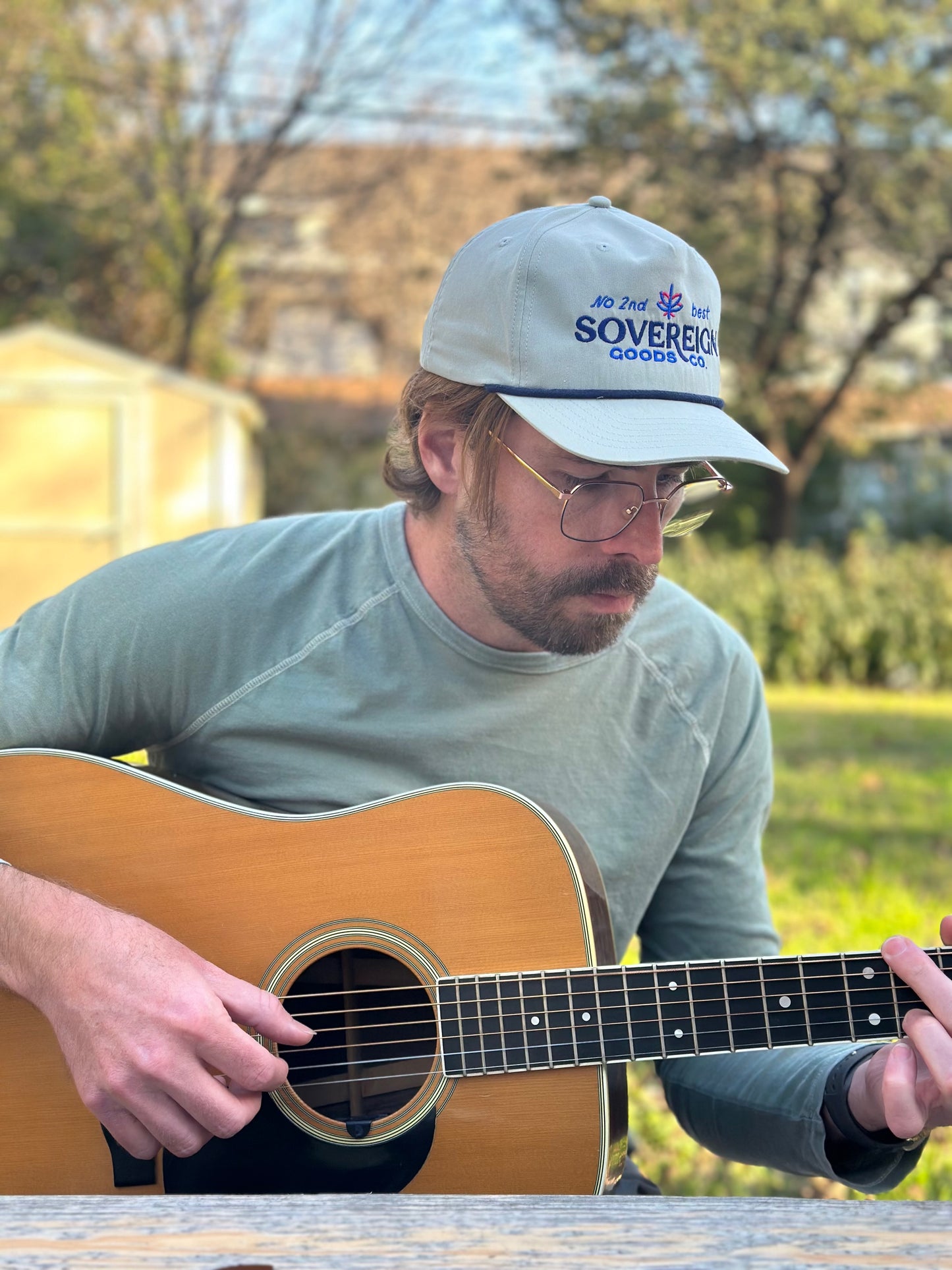 Man playing an acoustic guitar outdoors wearing a cap with 'Sovereign Goods' branding.