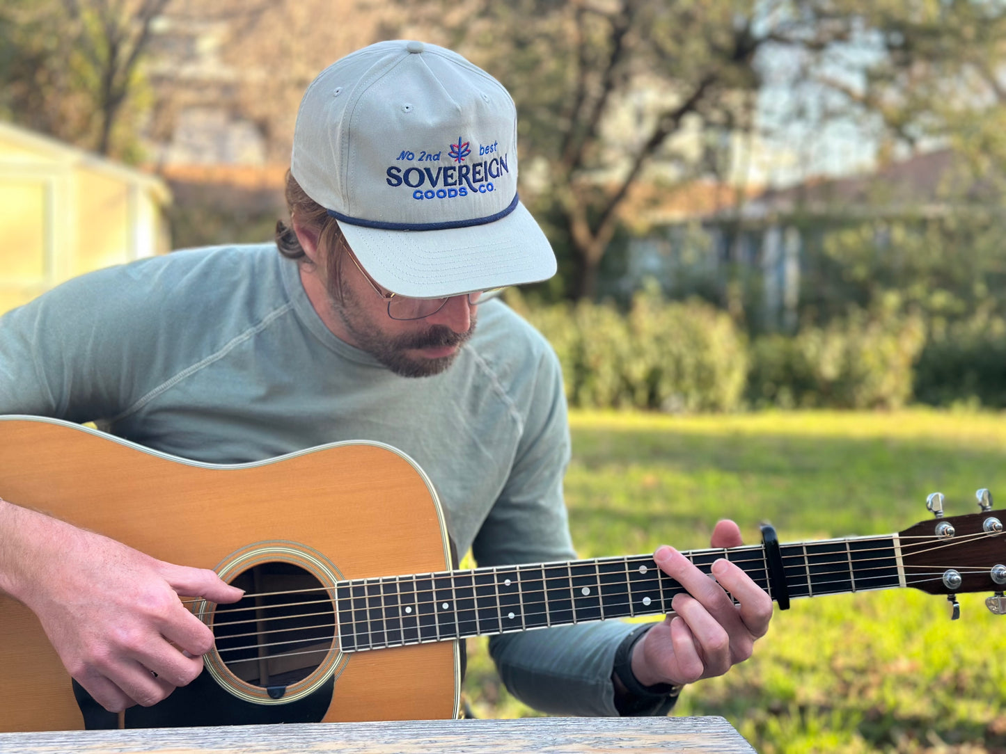 Man playing an acoustic guitar outdoors wearing a cap with 'Sovereign Goods' branding.