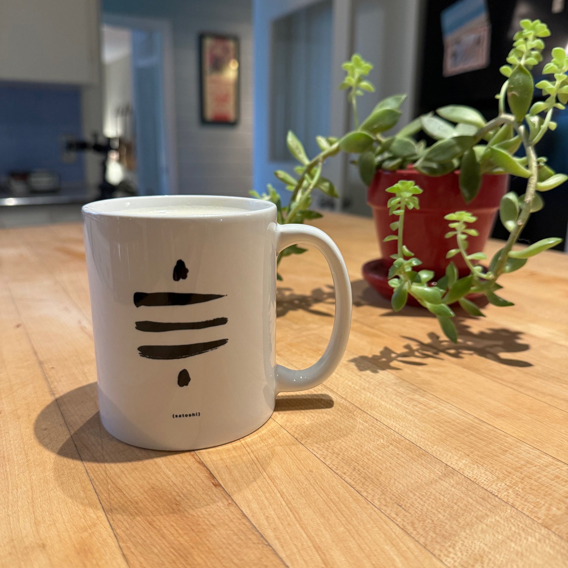 White Satoshi Symbol Mug with a black Sat Symbol and the text '(satoshi)' sitting on a butcher block counter with a green succulent