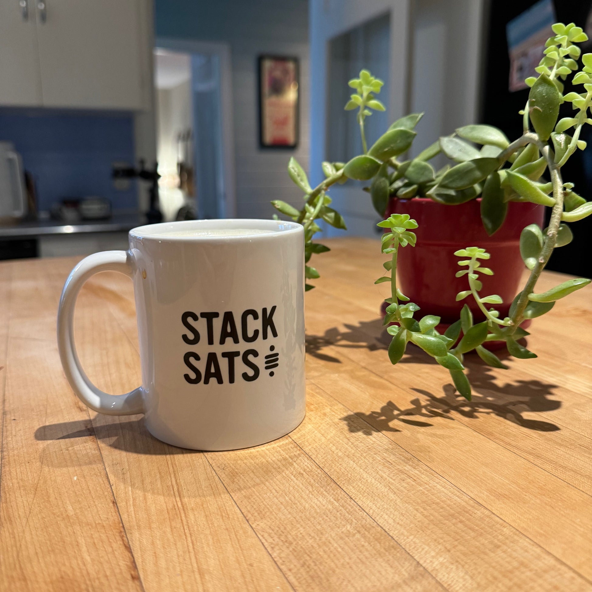 White Satoshi Symbol Mug with a black Sat Symbol and the text 'STACK SATS' sitting on a butcher block counter with a green plant