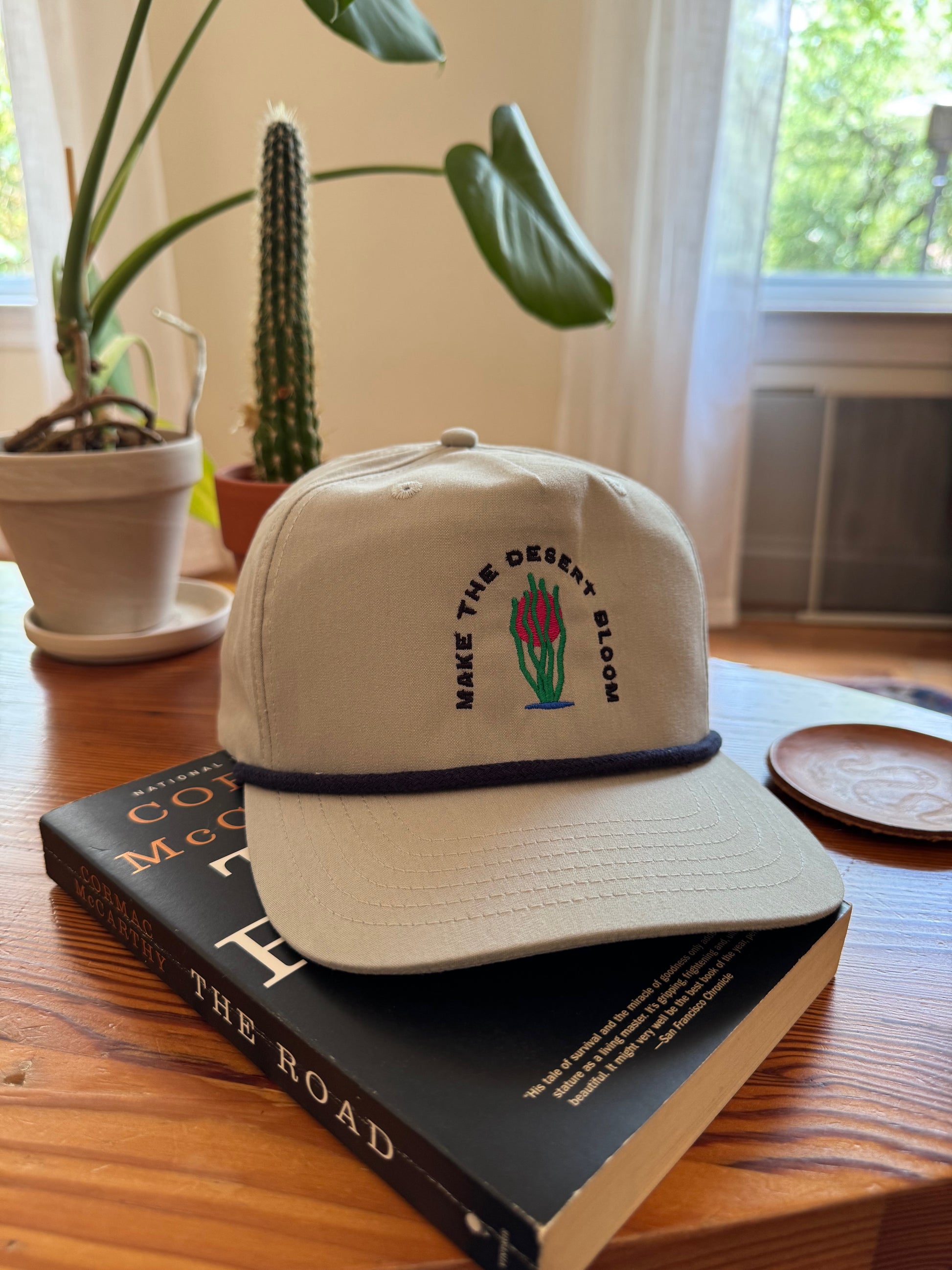 A grey golf rope cap with a navy rope sits on a wooden coffee table on top of a book called "The Road." Embroidered on the hat is an ocotillo in front of a setting sun and under the words "Make the desert bloom."