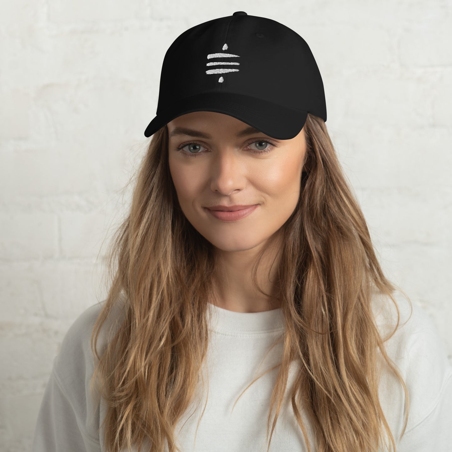 A female model with long blonde hair a beard wears a black dad hat with the Sat Symbol embroidered in white on the front.
